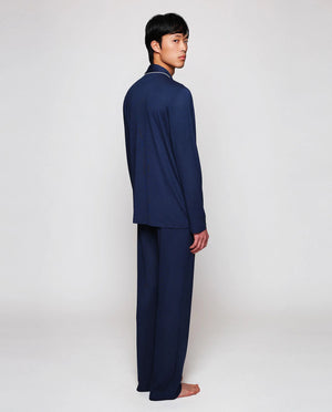 A person with short dark hair stands barefoot on a white background, wearing the MIRTO 1956 - NAVY MODAL PAJAMAS: a breathable matching set with white piping and buttons by MIRTO 1956.