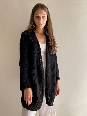 A person with long, brown hair is facing the camera, wearing a black KYLA CARDIGAN by DEBBIE KATZ over a white top and white pants. The background is plain and light-colored. They have a neutral expression and natural makeup.