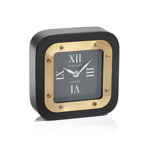 Two MARK LANE TABLE CLOCK - LARGE by ZODAX, square in shape with black and gold iron finish and Roman numerals, are displayed—one on a table, the other atop "Rooms to Inspire" books. The clocks show different times.