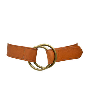 An ADA - JOSIE BELT in tan leather, featuring a double brass ring buckle, is displayed on a light blue surface. The belt, part of the ADA Collection, is coiled into an elongated spiral with one end draped over a cylindrical object of matching light blue color.