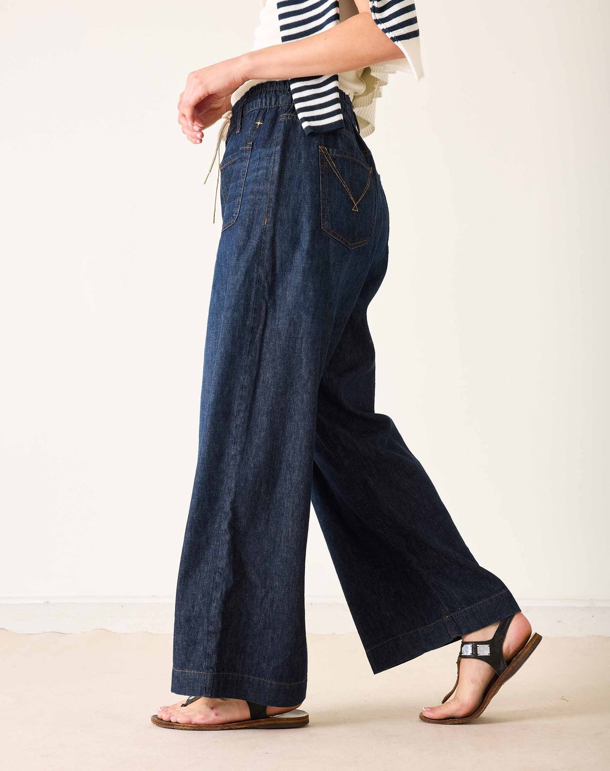 Wearing the MERSEA Londonberry Drawstring Jeans in Night Sky—a pair of wide-leg dark denim jeans with a comfortable drawstring waist—from MERSEA, styled with a cream sleeveless top, a striped shirt draped over the shoulders, and brown sandals, standing against a plain light background.