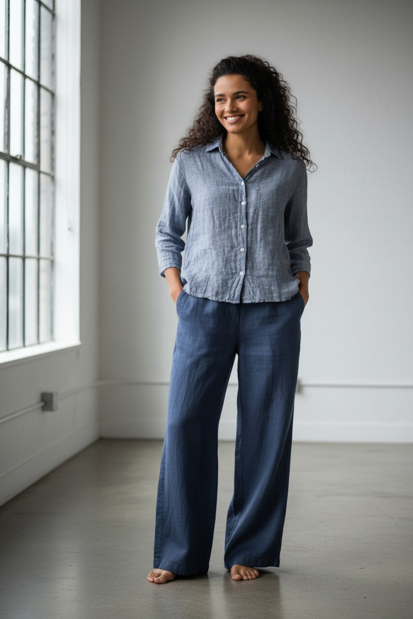 The CP SHADES - TESS LINEN WIDE LEG PANT by CP SHADES hangs on a black hanger against a light gray wall. These linen pants offer a relaxed fit with pockets, visible seams, and a slightly wrinkled texture.