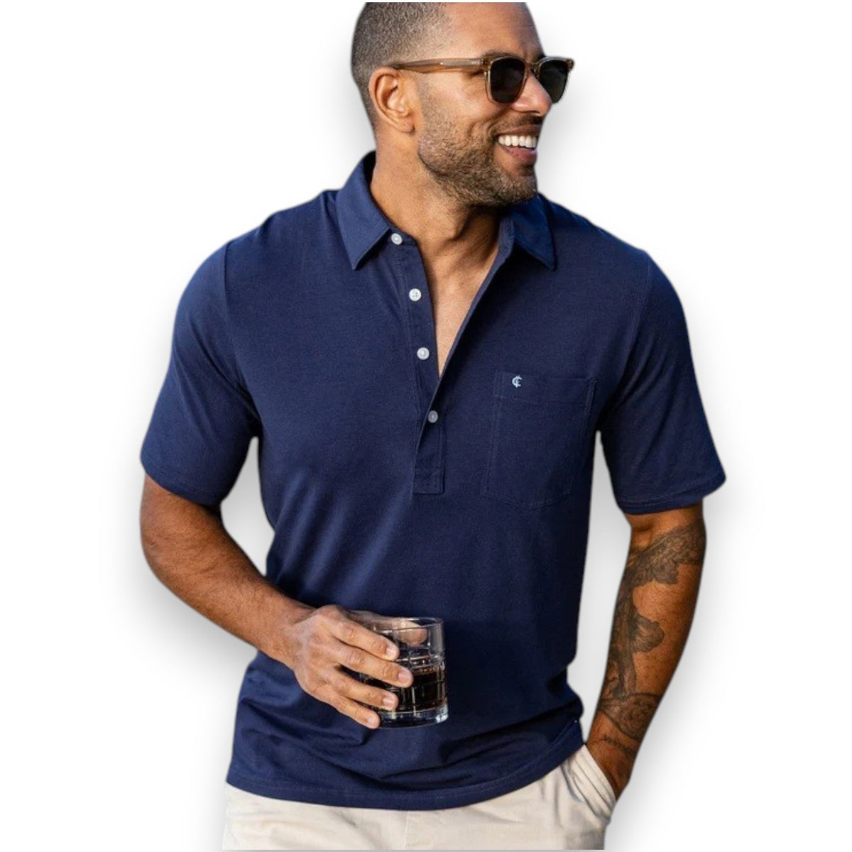 A smiling man in sunglasses stands outside by a wooden building, holding a drink and wearing the CRIQUET Top-Shelf Players Shirt in Navy. Sunlight and greenery are visible in the background.