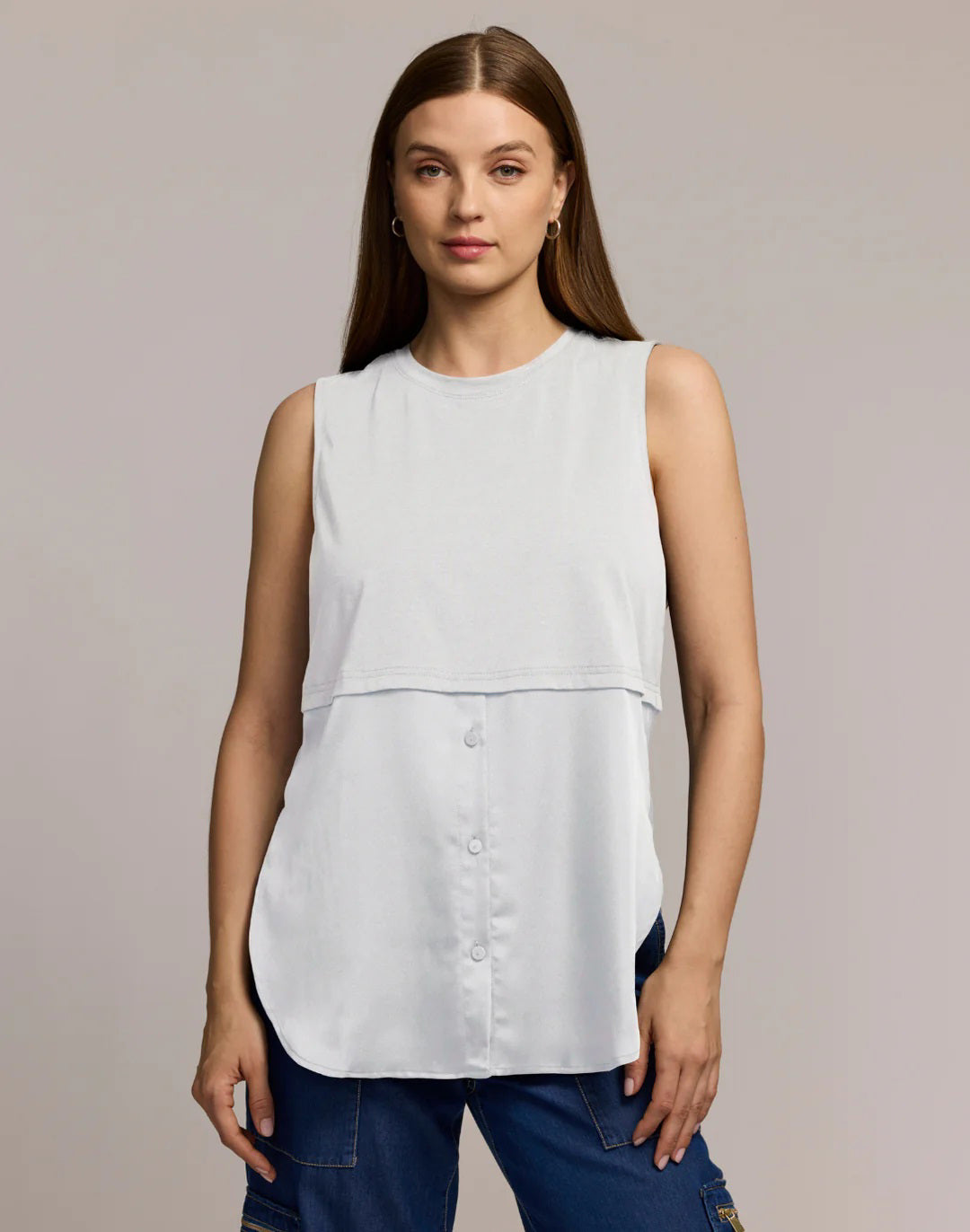 A woman with long brown hair wears the HINSON WU REMY Sleeveless Foundation Layer Shirt in white and blue jeans, standing against a light gray background. She faces forward with a neutral expression, her left hand by her side.