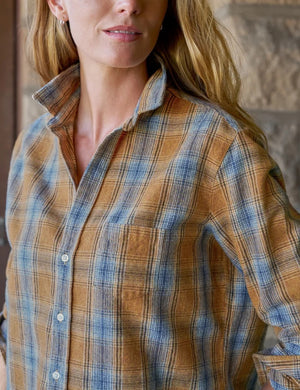 A woman stands indoors against a neutral background with soft light streaming in, smiling gently with one hand in her pocket. She wears the FRANK & EILEEN Eileen Relaxed Button Up Shirt in Italian Flannel Gold Blue Large Plaid—featuring a bust-flattering button placement—paired with cream high-waisted pants and brown ankle boots.