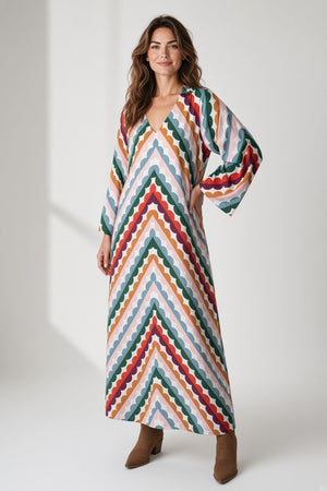 A woman wears a BRITON COURT MILANI MAXI DRESS IN GROOVY GROVE, a long-sleeved, v-neck maxi dress with a multicolored scalloped chevron print and brown ankle boots.