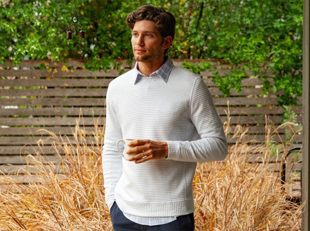 A smiling man with a short beard wears the CRIQUET - QUILTED CREW SWEATSHIRT IN OFF-WHITE by CRIQUET, holding a green can indoors near wood-paneled walls, a window, and a wooden chair.
