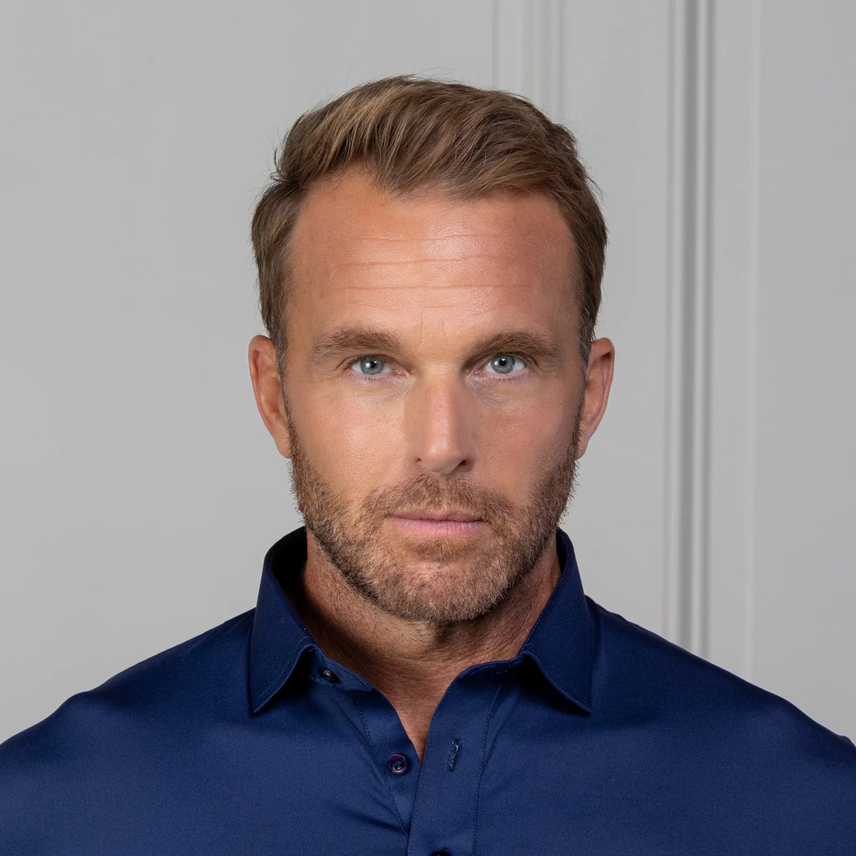 A man in the COLLARS AND CO FEATHERWEIGHT FLEX POLO IN NAVY and white pants stands by a light-paneled wall, smiling and holding the side of his performance golf shirt.
