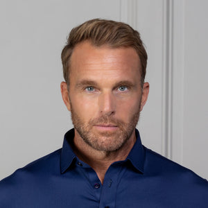A man in the COLLARS AND CO FEATHERWEIGHT FLEX POLO IN NAVY and white pants stands by a light-paneled wall, smiling and holding the side of his performance golf shirt.