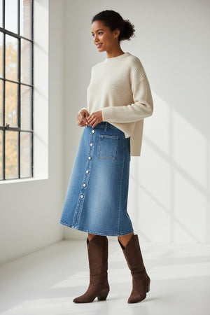A woman walks on a dirt road near a fence, wearing a tan button-up shirt and the FRANK & EILEEN - SHANNON CLASSIC DENIM SKIRT IN 1978 WASH, paired with brown boots. In the background, trees and horses stand in a grassy field.