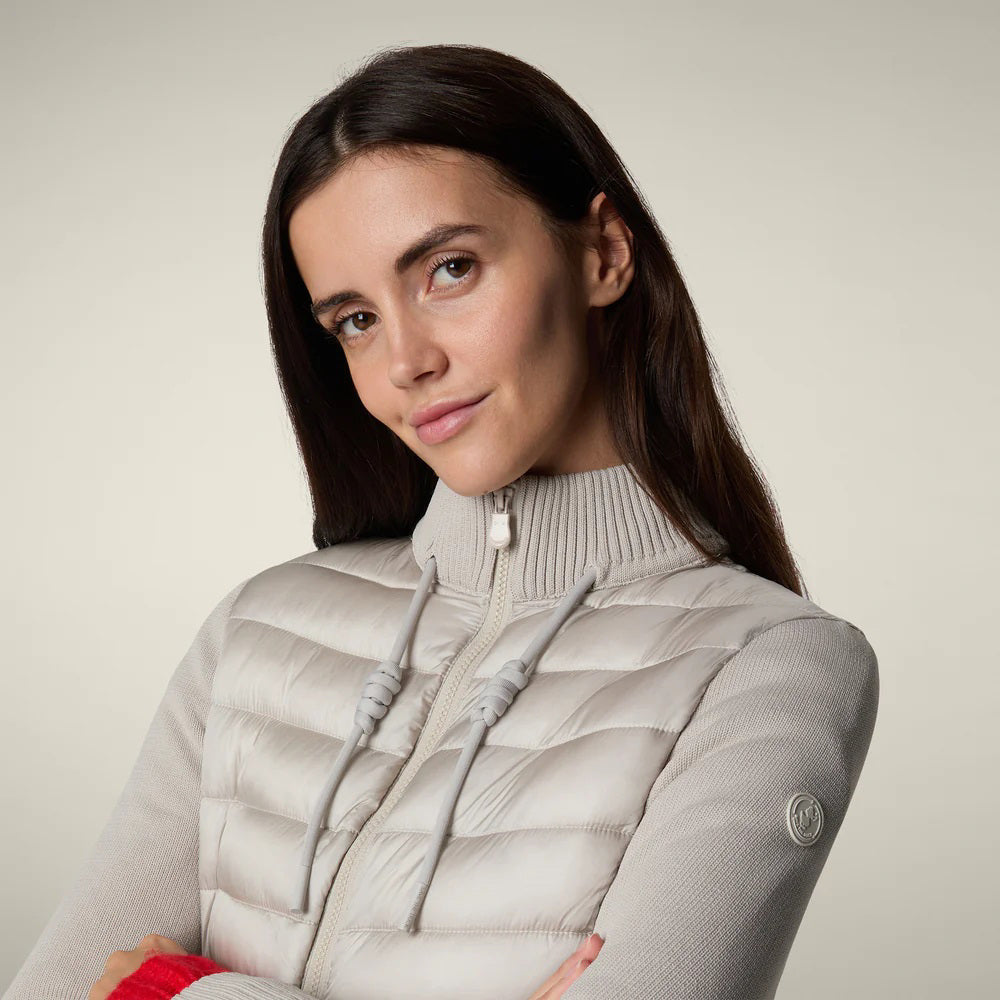 A woman with straight brown hair and a neutral expression models the SAVE THE DUCK - ZILIA JACKET in light gray, styled with olive cargo pants, red socks, and tan boots—showcasing effortless transitional layering.