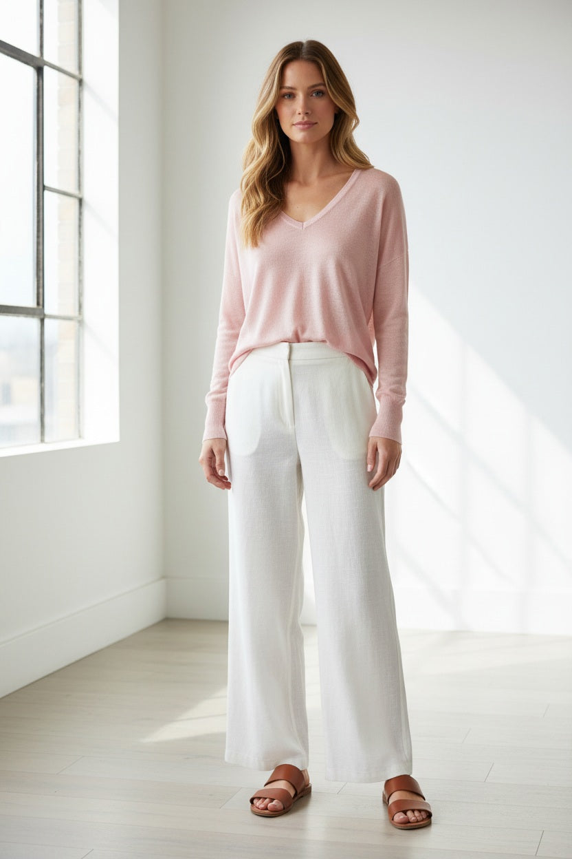 A woman smiles with one hand on her hip against a white background, wearing a KINROSS CASHMERE Easy Vee Pullover in Linen paired with white wide-leg pants, sandals, and her long hair down.