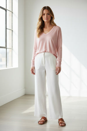 A woman smiles with one hand on her hip against a white background, wearing a KINROSS CASHMERE Easy Vee Pullover in Linen paired with white wide-leg pants, sandals, and her long hair down.