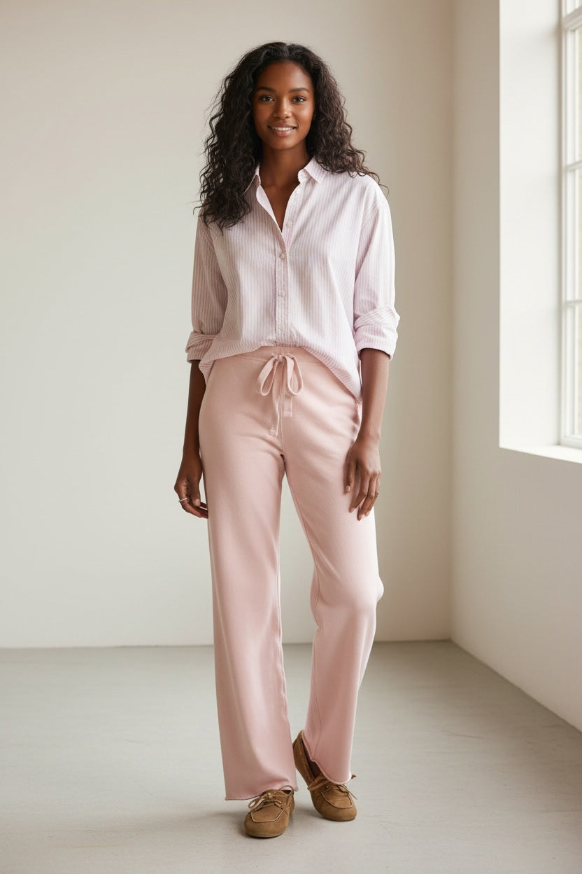 Clad in the FRANK &amp; EILEEN Catherine Long Sweatpant in Triple Fleece Antique Pink, a person stands in a cozy living room with neutral decor and wooden furniture, paired with a light-colored striped shirt and brown moccasin slippers.