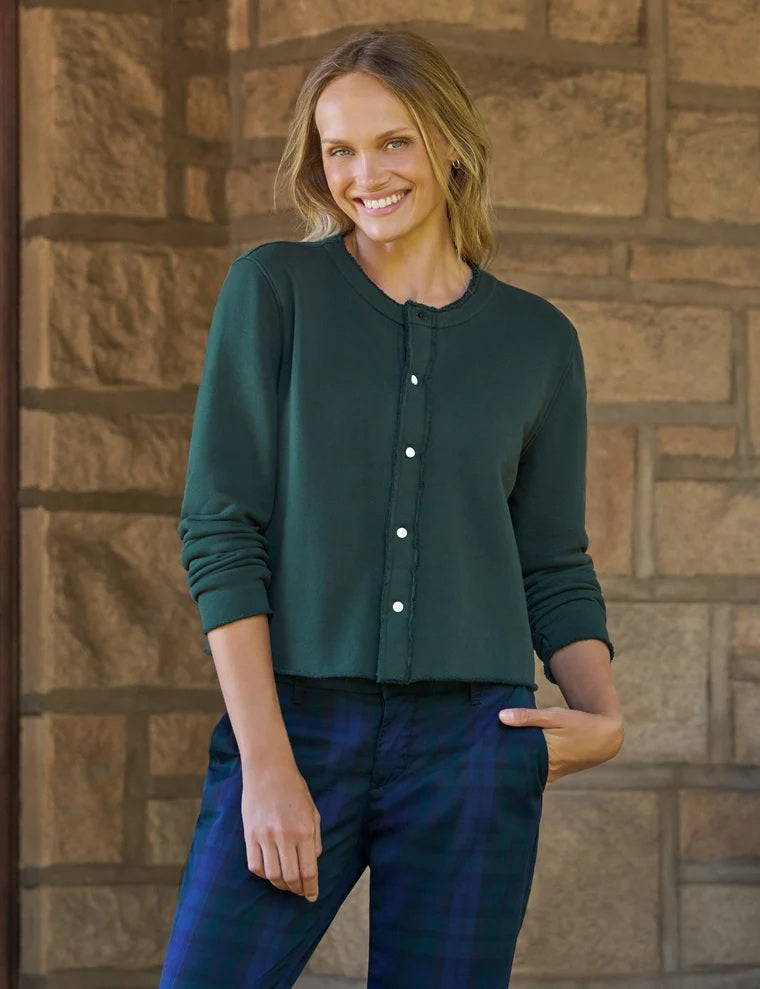 A woman with wavy dark hair stands by large windows, smiling as she wears the FRANK & EILEEN Jackie Sweatshirt Cardigan in Triple Fleece Pine from FRANK & EILEEN, paired with blue plaid pants and black loafers. One hand is tucked into her pocket and her legs are crossed.