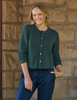 A woman with wavy dark hair stands by large windows, smiling as she wears the FRANK & EILEEN Jackie Sweatshirt Cardigan in Triple Fleece Pine from FRANK & EILEEN, paired with blue plaid pants and black loafers. One hand is tucked into her pocket and her legs are crossed.
