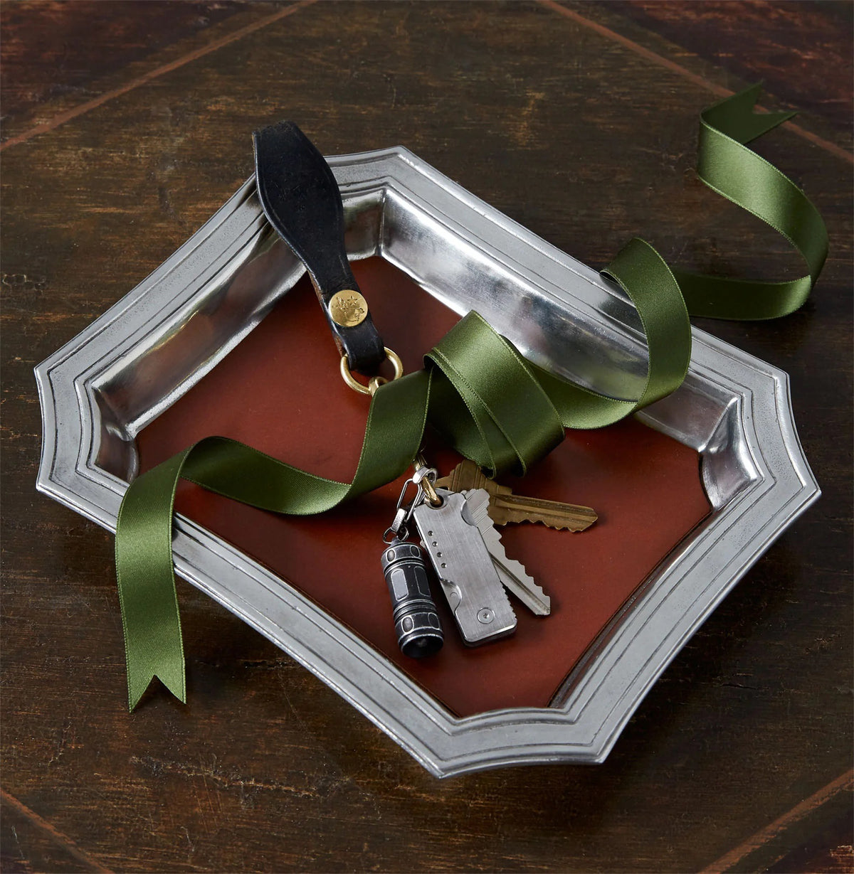 The MATCH - PEWTER POCKET CHANGE TRAY by MATCH, featuring a dark brown base, holds a black leather keychain, three keys, and a small flashlight charm wrapped in green satin ribbon. The tray sits on a dark wood surface.