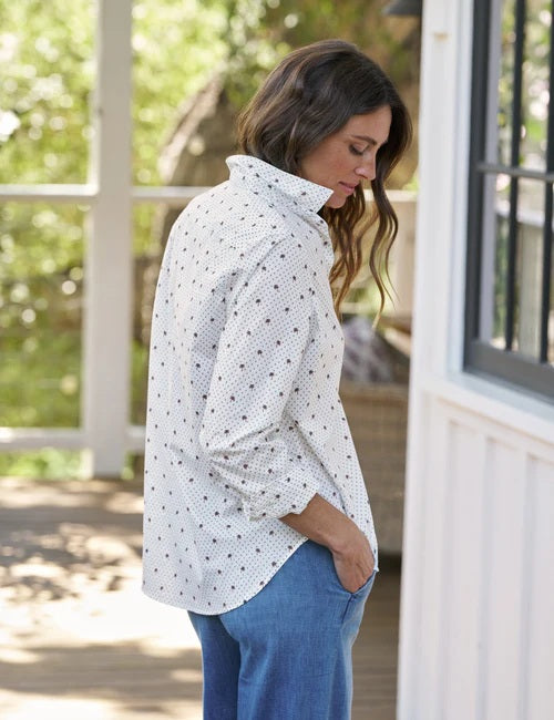 A woman wearing the FRANK & EILEEN Eileen Relaxed Button Up in Italian Cotton Palm Trees paired with blue jeans stands outside by a white building with large windows, smiling softly with one arm resting on her hip; the flattering button placement of the shirt by FRANK & EILEEN accentuates her silhouette.