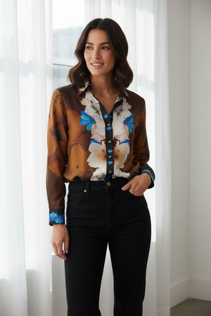 A woman stands indoors by a window, smiling as she looks to the side. She wears the FRANCO FERRARI Long Sleeve Silk Blouse, featuring a vibrant floral print in brown, white, and blue patterns, paired with black high-waisted pants. One hand is in her pocket.