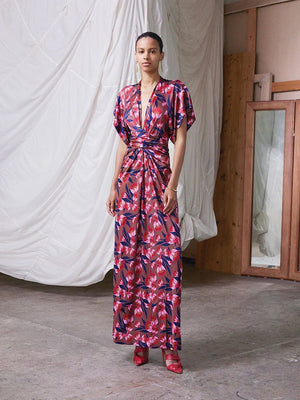 A woman stands indoors wearing the FRANCO FERRARI - JERSEY PRINT DRESS, a long, deep V-neck jersey dress with short sleeves and a red and blue botanical print. She pairs it with red shoes against draped white fabric and wooden frames.