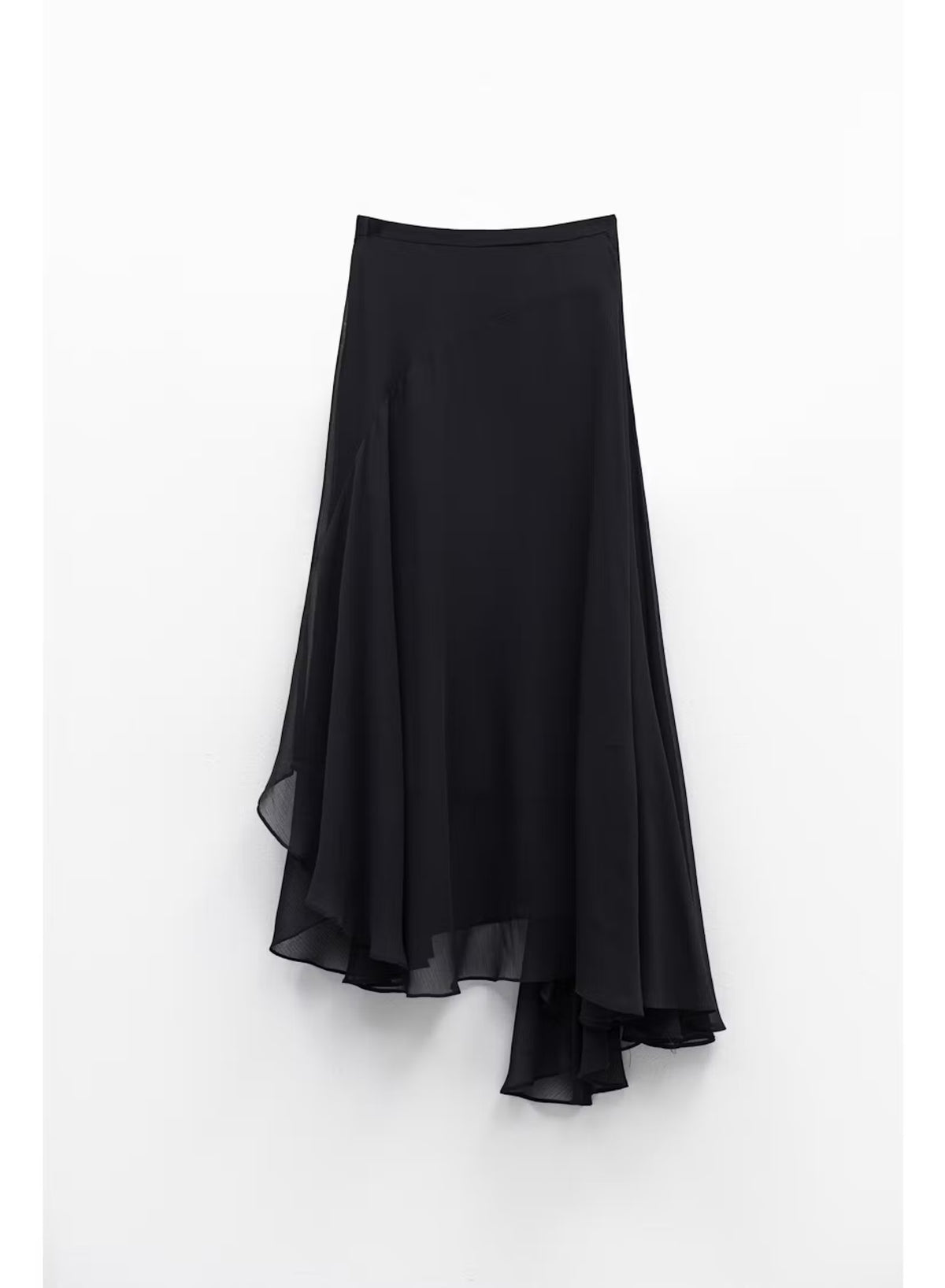 A woman stands indoors by large windows, showcasing a full-length silhouette in the ELEH Asymmetrical Skirt in Black, paired with a black short-sleeve top and black ankle boots.