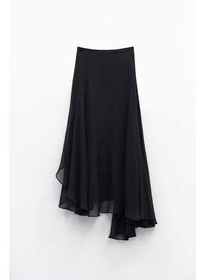 A woman stands indoors by large windows, showcasing a full-length silhouette in the ELEH Asymmetrical Skirt in Black, paired with a black short-sleeve top and black ankle boots.