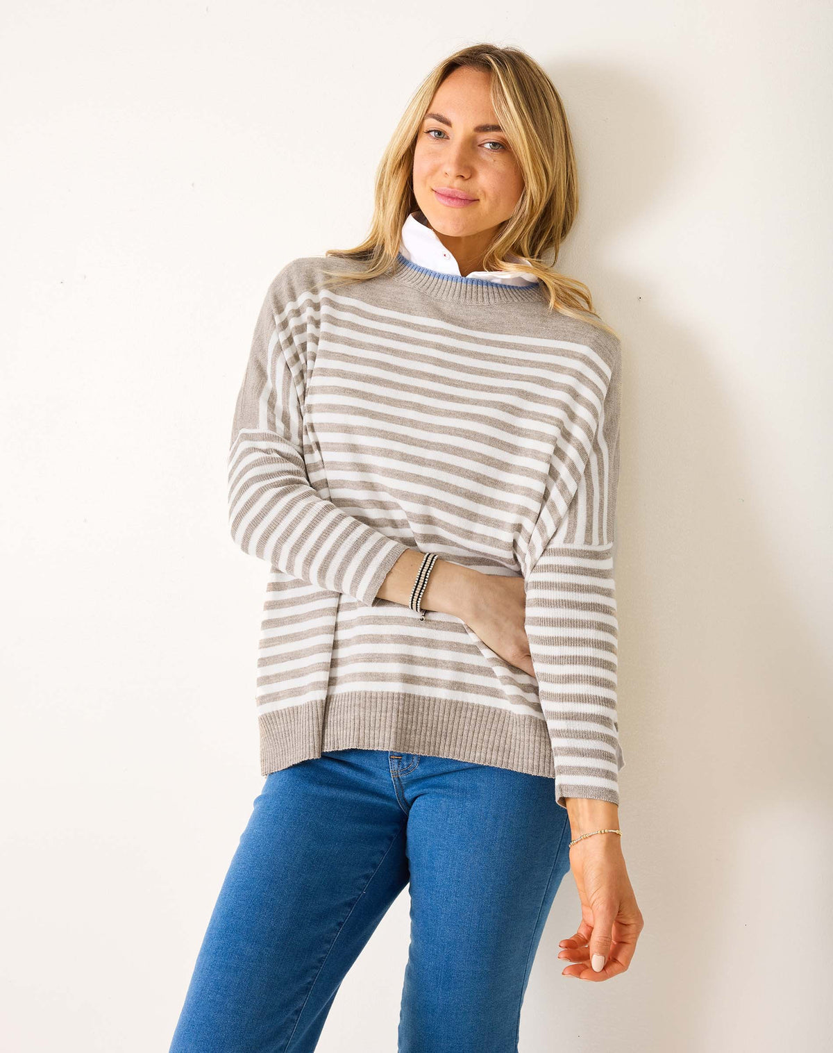 A woman with blonde hair wears the MERSEA - CATALINA MODERN CREWNECK SWEATER in gray and white stripes over a white collared shirt and blue jeans, standing against a plain white wall with a slight smile, one arm bent across her body.