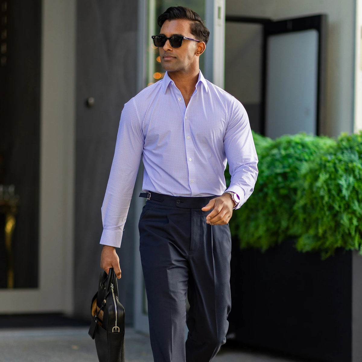 A man in COLLARS AND CO's Semi Spread Collar Apex Performance Dress Shirt in Lavender and Blue Multi Gingham adjusts his collar outdoors, surrounded by green plants and a blurred background.