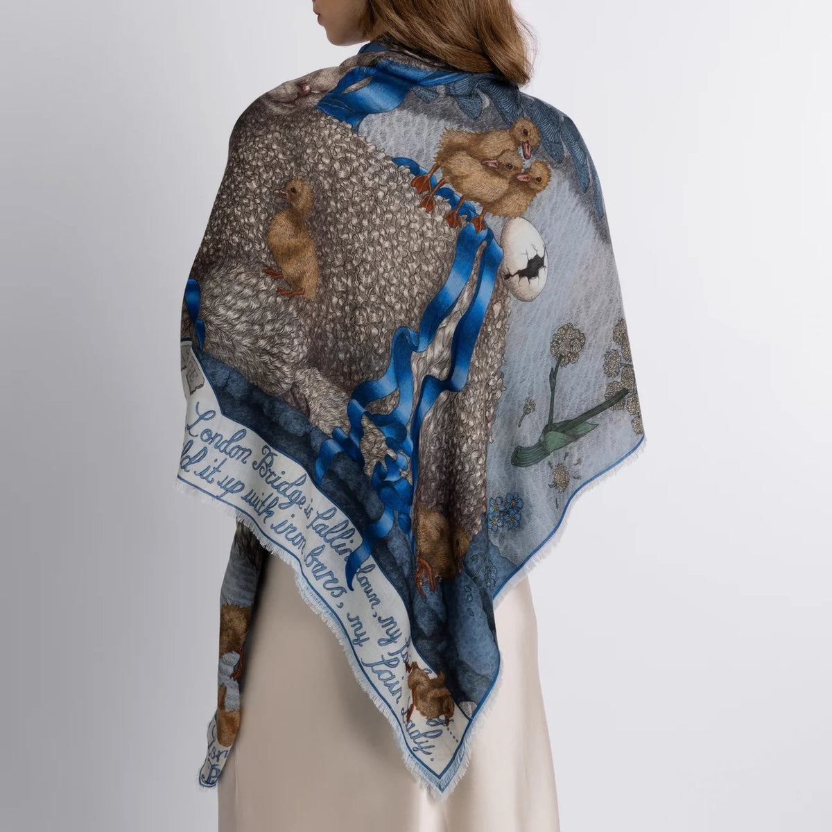 The SABINA SAVAGE ENGLAND cashmere scarf, named "London Bridge is Falling Down," displays intricate nature-themed illustrations of rabbits, flowers, and baby lambs on a light blue background. Tied in a loose knot, the scarf beautifully highlights its detailed patterns.