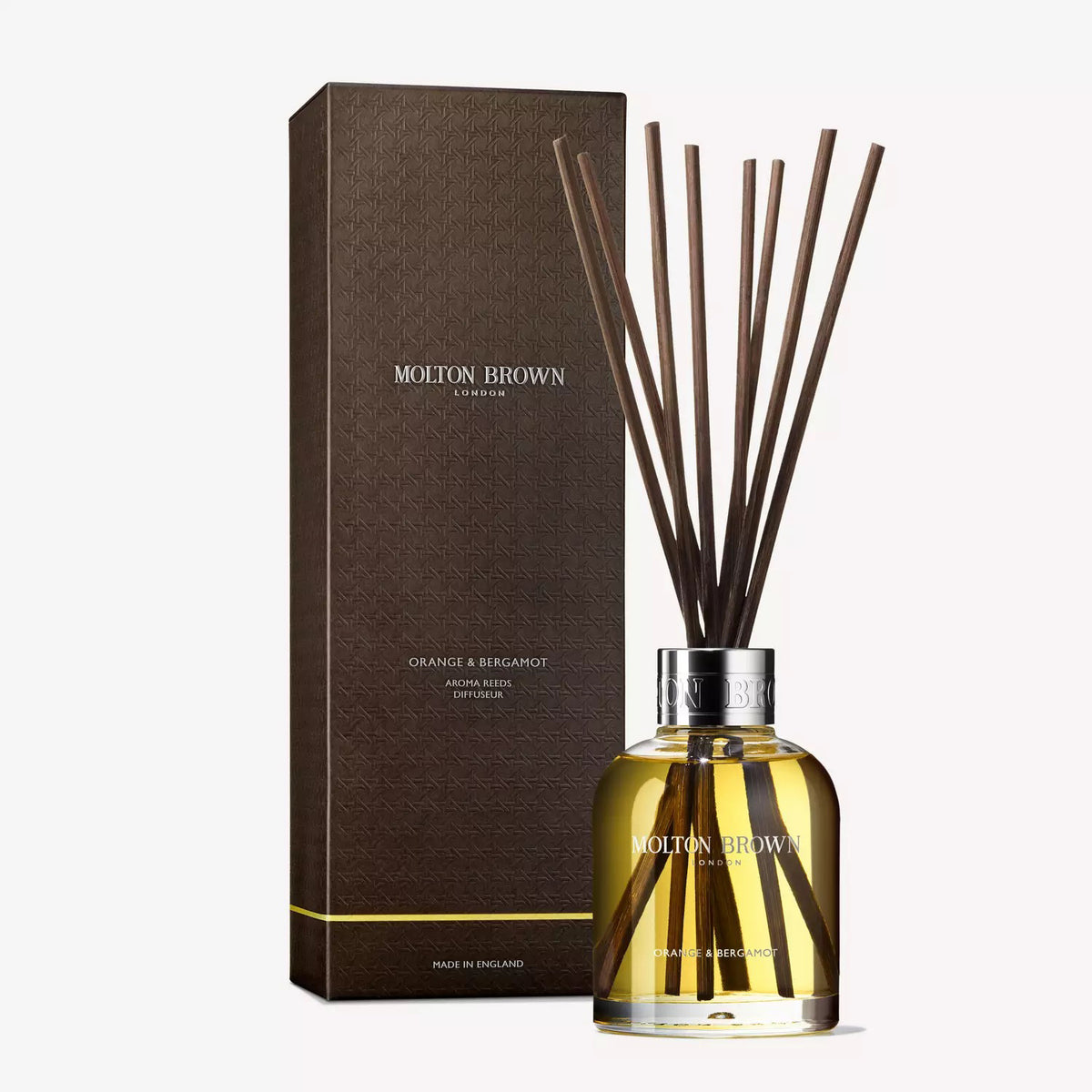 A clear glass bottle filled with yellow liquid, labeled &quot;MOLTON BROWN - ORANGE AND BERGAMOT DIFFUSER,&quot; features several long, thin, dark brown diffuser reeds extending from its neck. The reed diffuser boasts a hint of Sevillian orange and neroli. A metallic ring at its opening gives it an elegant and classy appearance.