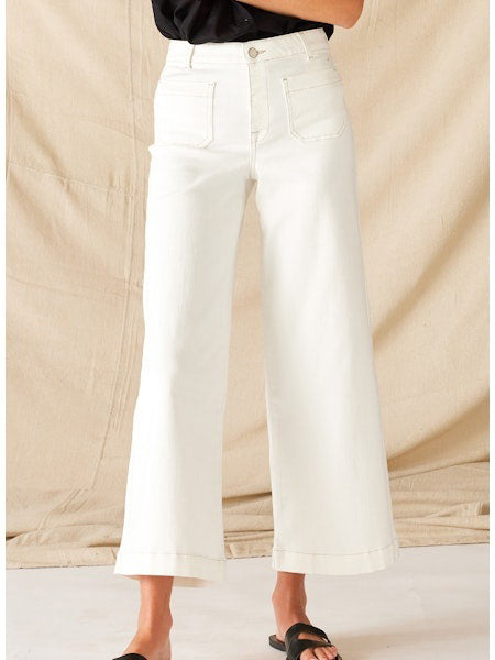 Someone wearing the MERSEA COCO SAILOR WIDE LEG DENIM PANT in white, featuring high-waisted, nautical-inspired style with front patch pockets, paired with black sandals and standing against a beige fabric backdrop.
