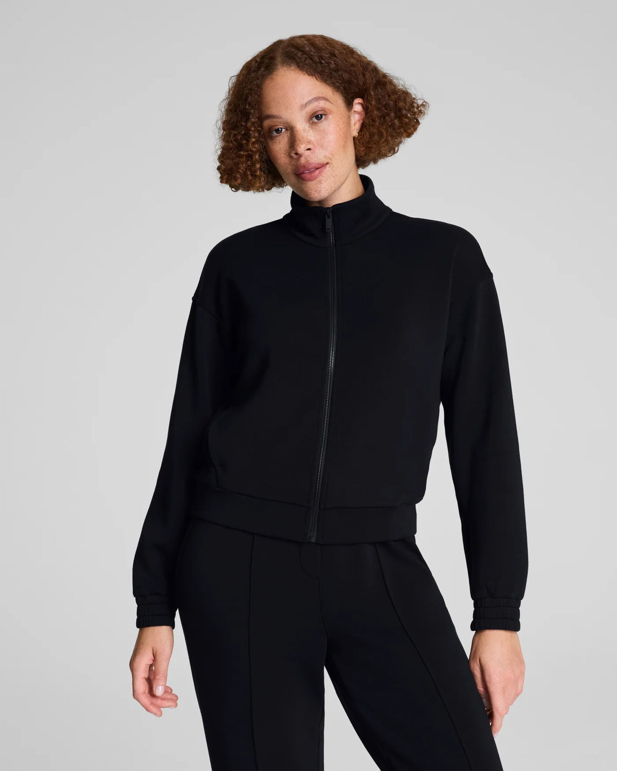 A person with curly hair models the SPANX AIRESSENTIALS BRUSHED FULL ZIP JACKET in black with matching pants, standing against a light gray background—highlighting chic loungewear from SPANX.