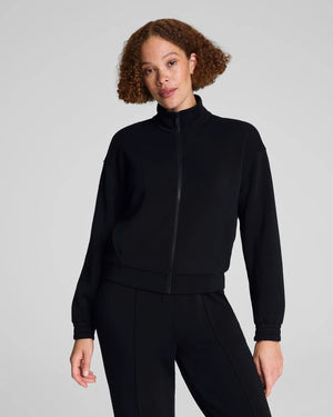 A person with curly hair models the SPANX AIRESSENTIALS BRUSHED FULL ZIP JACKET in black with matching pants, standing against a light gray background—highlighting chic loungewear from SPANX.
