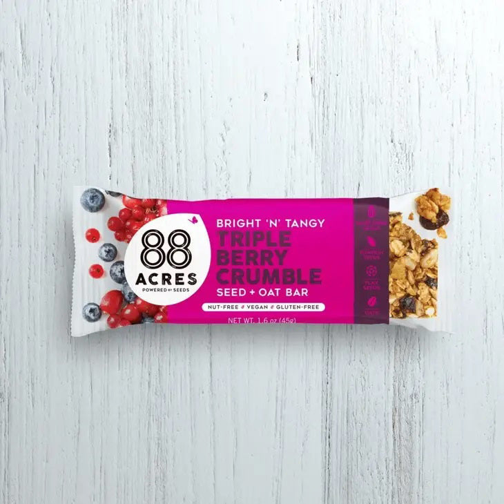 An 88 ACRES - TRIPPLE BERRY CRUMBLE Seed + Oat Bar rests on a light wooden surface, with its package partially open to show a portion of the nut-free and vegan granola bar inside. The packaging is white, featuring a vibrant purple section that displays the product name and various icons.