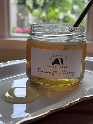 The Fearrington Signature Collection's hexagonal glass jar elegantly holds their Wildflower Honey, known for its raw quality. The label states: "Raw, Unfiltered. Handmade with care in our kitchens. May contain allergens. 282 grams, 10 oz." Inside is the finest golden honey from Fearrington House.