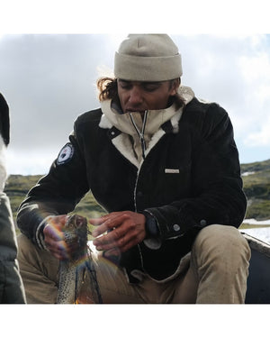 A person in a warm jacket and beanie smiles while holding a red-feathered bird, possibly on a boat, wearing the AMUNDSEN SPORTS - HARVESTER OVERSHIRT.