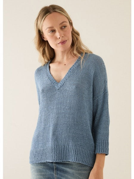 A smiling woman with long blonde hair wears the BASE MILANO - V NECK COTTON SWEATER, a light blue, short-sleeved, relaxed fit knit by BASE MILANO, paired with blue jeans. She stands against a plain background, hand on her chin.