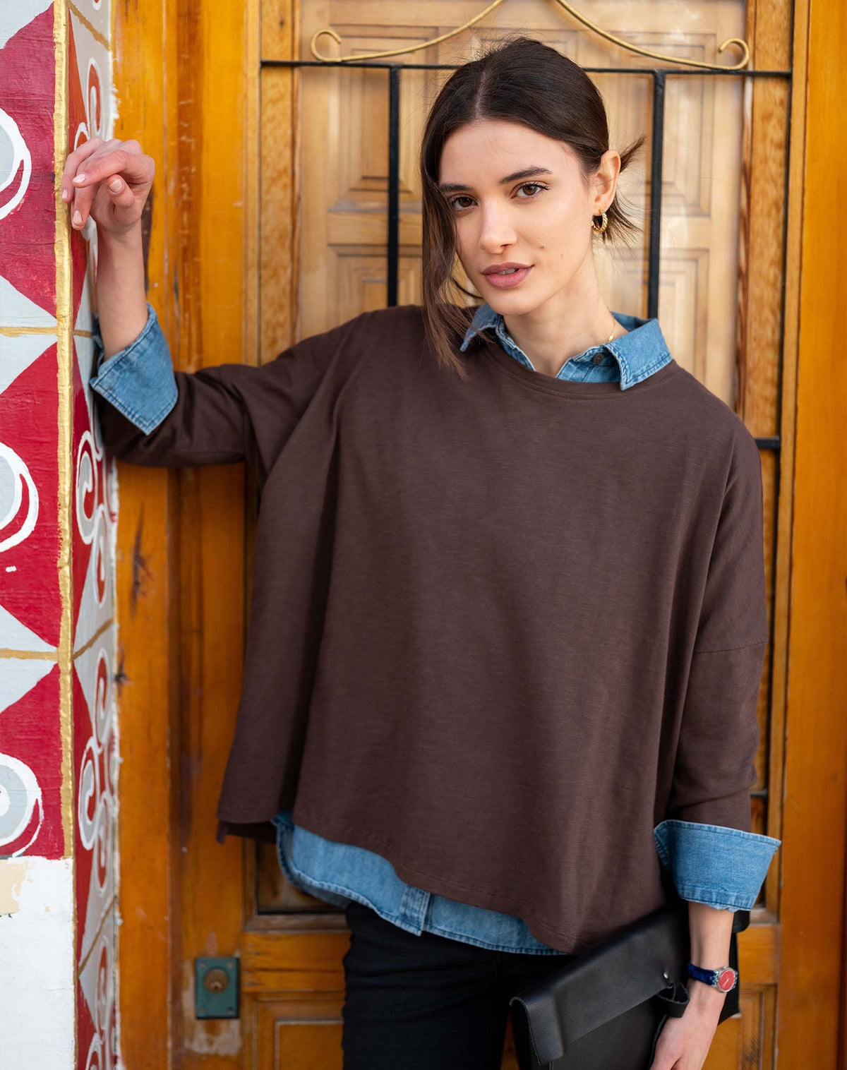A woman with dark hair stands by a wooden door, wearing the MERSEA - Catalina Slub Tee in Espresso layered over a blue denim shirt and holding a black clutch. She looks at the camera, resting her arm on a red and white tiled wall.
