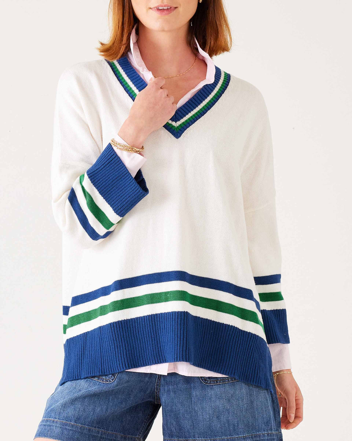 A woman wears the MERSEA - MONTAUK V-NECK SWEATER by MERSEA, a white oversized soft knit with blue and green striped collar, cuffs, and hem, ribbed v-neckline, styled over denim shorts and a light pink shirt.