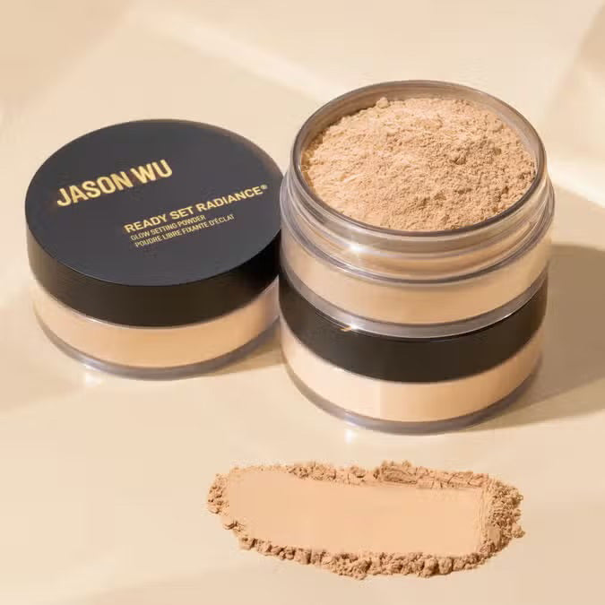 An open container of JASON WU - READY SET RADIANCE POWDER by JASON WU BEAUTY displays its light beige contents. Beside it, a closed container and a smudged swatch of the powder, which promises a luminous finish enhanced with Vitamin C, rest on a light-colored surface.