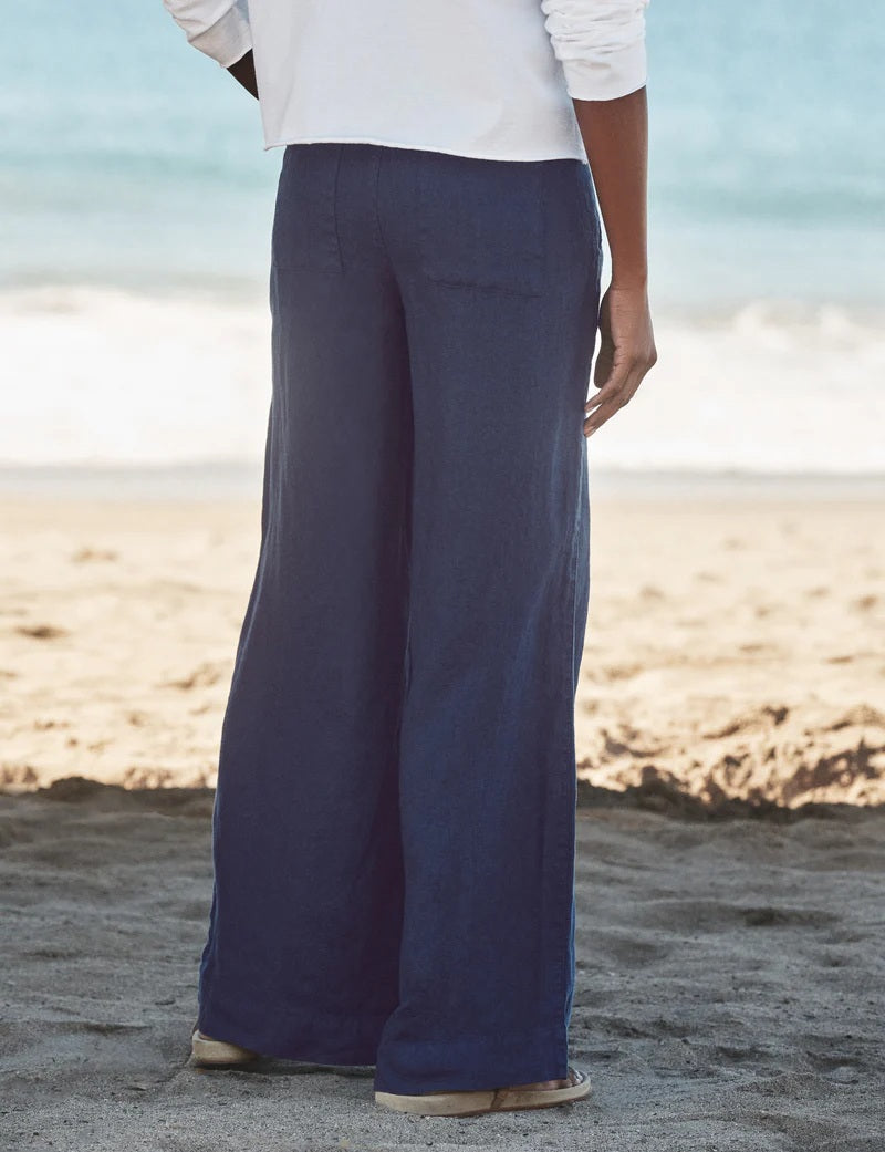 A person stands on a sandy beach wearing a white long-sleeve shirt, FRANK &amp; EILEEN MADELINE Patch Pocket Pull On Wide Leg Linen Pant in Navy, and light-colored sandals. The ocean is visible in the background.