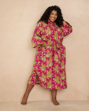 A person stands barefoot against a light wall, wearing the PRINTFRESH - BAGHEERA COTTON ROBE. This colorful floral-patterned robe, crafted from organic cotton poplin and hand screen-printed in vibrant hot pink hues, ties at the waist and falls below the knees. The person has long hair and smiles softly.