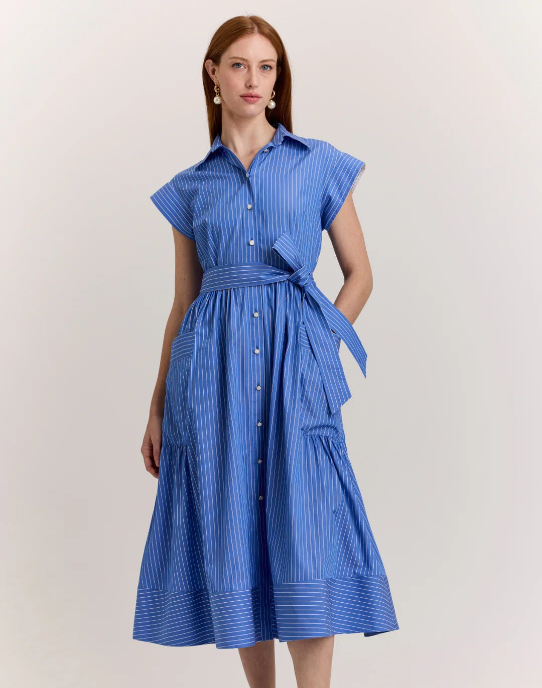 A woman with long brown hair models the HINSON WU Annabelle Short Sleeve Dress in Lake Blue Pencil Stripe, featuring a collared, button-down 100% cotton design with a matching waist tie. She stands against a plain light background.