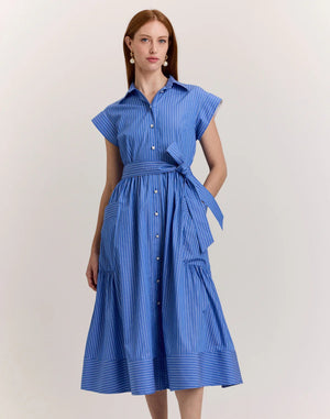 A woman with long brown hair models the HINSON WU Annabelle Short Sleeve Dress in Lake Blue Pencil Stripe, featuring a collared, button-down 100% cotton design with a matching waist tie. She stands against a plain light background.