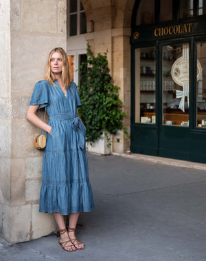 A woman with blonde hair, wearing the MERSEA Pariscope Maxi Dress in Chambray, stands by a stone wall. The blue denim maxi dress from MERSEA features short sleeves, a belted waist, and a subtle ruffle neckline for added elegance. She accessorizes with a gold necklace featuring a round pendant.