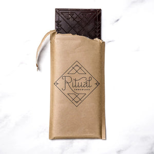 A green RITUAL CHOCOLATE - FLEUR DE SEL CHOCOLATE BAR 70% CACAO with silver geometric designs and text on the wrapper, shown on a white marble background.