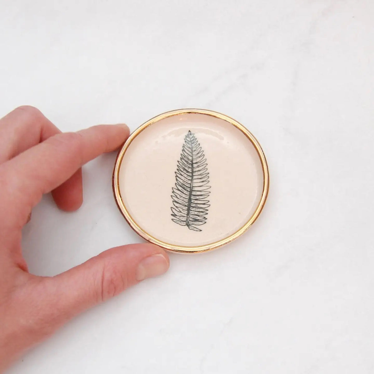 The COMPOST DESIGN - FERN TINY DISH 3&quot; features a black fern leaf on white stoneware, highlighted by a 22k gold rim. Its light, neutral tones and hand-built rustic look beautifully contrast against the white marble surface.
