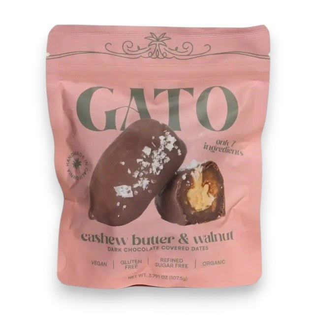A pink pouch of GATO DATES - CHOCOLATE COVERED CASHEW BUTTER & WALNUT DATES by GATO DATES, featuring an image of two chocolate pieces—one sliced open to reveal the filling. The packaging highlights that it’s vegan, gluten-free, organic, and proudly plant-based.