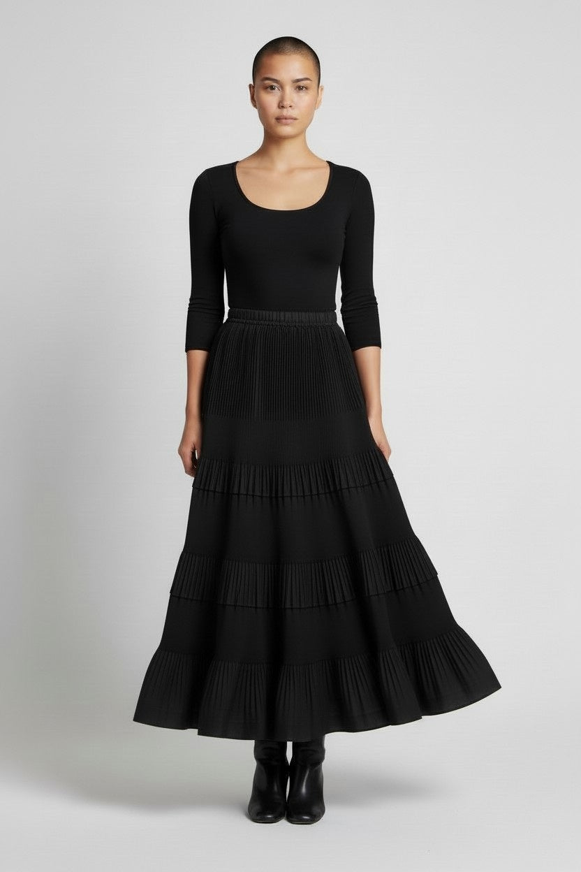 A person with short hair stands against a plain background wearing a fitted black long-sleeve top, MEIMEIJ’s RUFFLE SKIRT IN BLACK falling to the ankles, and black ankle boots.