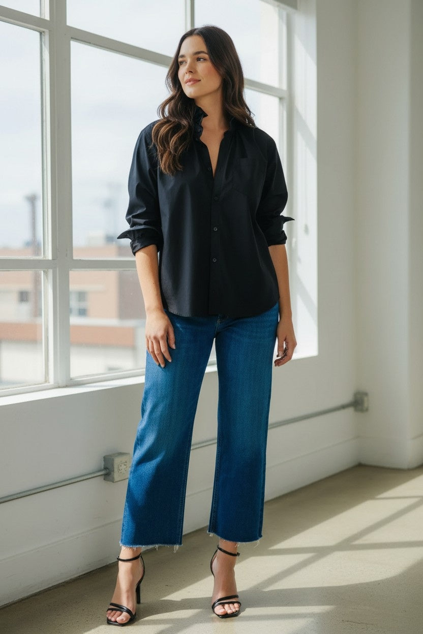 A woman with wavy dark hair and red lipstick leans against a wall outdoors, smiling at the camera. She wears the KMJ - QUARANTINA SHIRT by KMJ, sequined green pants, and gold jewelry. Green foliage and a sidewalk are behind her.